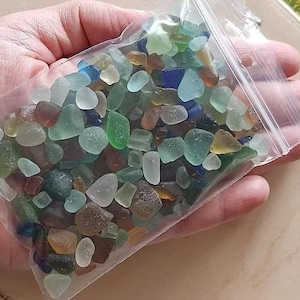May include: A clear plastic bag filled with a variety of small, colorful, and iridescent sea glass pieces. The sea glass is a mix of blues, greens, browns, and clear glass.