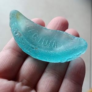 Puede incluir: Un trozo de vidrio de mar azul claro en forma de media luna con la palabra "WIUM" grabada en él.