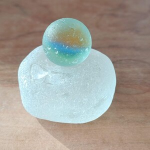 Puede incluir: Un pequeño mármol redondo de vidrio verde y azul con un diseño de remolino blanco y naranja se asienta sobre una roca de vidrio blanco mate lisa.