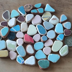 Può includere: Una collezione di piccoli pezzi di ceramica di forma irregolare in vari colori pastello, tra cui blu, rosa e verde, con una moneta per la scala. I pezzi hanno un aspetto liscio e arrotondato e sono disposti su una superficie di legno.