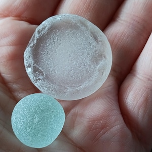 Puede incluir: Dos piezas de vidrio de mar, una transparente y otra turquesa, descansan en una palma abierta. La pieza más grande tiene una forma redondeada e irregular. La más pequeña es lisa y redonda. Ambas tienen un aspecto esmerilado.