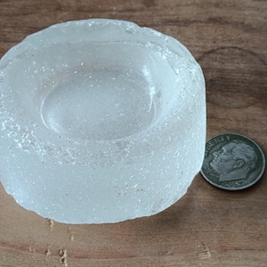 Puede incluir: Un portavelas votivo redondo de vidrio transparente y texturizado. Una moneda está al lado del portavelas. El portavelas mide aproximadamente 4 cm de diámetro y 2,5 cm de alto.