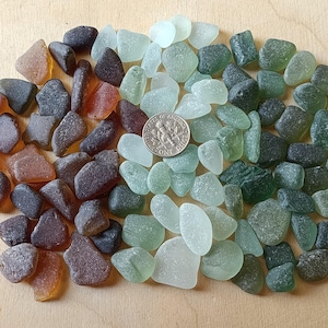 May include: A collection of smooth, weathered sea glass pieces in shades of brown, amber, green, and pale aqua. A silver-colored United States dime coin is placed among the glass fragments to provide a sense of scale for the various sizes.