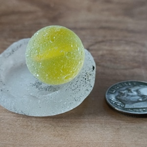 Puede incluir: Una canica de vidrio amarillo-verdoso se encuentra sobre un pequeño cuenco de vidrio marino transparente. Una moneda de plata está al lado del cuenco. Los objetos están sobre una superficie de madera.