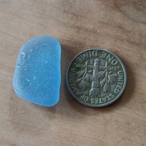 May include: A piece of smooth, light blue sea glass next to a United States dime coin. The sea glass is an irregular shape, and the coin shows the words "United States of America" and "One Dime".