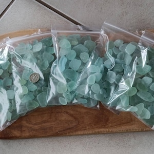 May include: Four clear plastic bags filled with sea glass in shades of blue and green. A coin is visible in one bag. The bags are arranged on a wooden cutting board.