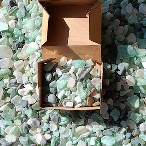 Puede incluir: Una caja de cartón llena de una variedad de vidrio de mar, incluyendo piezas verdes, azules y blancas. El vidrio de mar está esparcido alrededor de la caja, creando una exhibición colorida y texturizada.