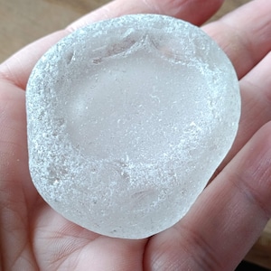 Puede incluir: Una piedra de vidrio de mar lisa y blanca con una textura escarchada. La piedra es redonda y tiene un centro ligeramente cóncavo.