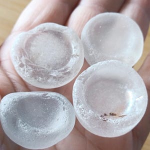 May include: Four pieces of sea glass in varying shapes and sizes, held in a hand. The glass is translucent with a frosted appearance, and the shapes are rounded and smooth. The sea glass is a pale, milky white color.