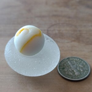 Puede incluir: Una canica blanca y amarilla se encuentra sobre una base de vidrio esmerilado, redonda y transparente, junto a una moneda de diez centavos estadounidense. La canica tiene un remolino amarillo y blanco. La moneda es de color plateado.
