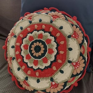 Könnte beinhalten: Ein rundes Häkelkissen mit einem mehrfarbigen Mandala-Muster. Das Kissen ist in verschiedenen Farben gearbeitet, darunter Rot, Orange, Grün und Beige.