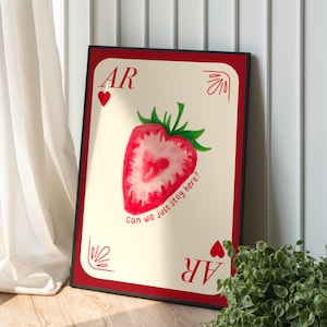 Puede incluir: Impresión artística enmarcada con un diseño de fresa en acuarela. La fresa roja tiene un centro en forma de corazón y hojas verdes. El texto incluye "AR" y "can we just stay here?" sobre un fondo crema con borde rojo.