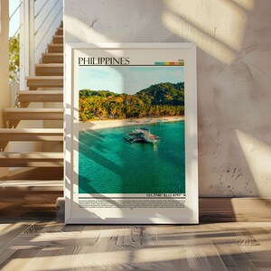 May include: White framed print featuring a tropical beach scene in the Philippines. The image shows a boat on turquoise water, a sandy beach, and lush green trees. The word "PHILIPPINES" is printed at the top, with coordinates at the bottom.