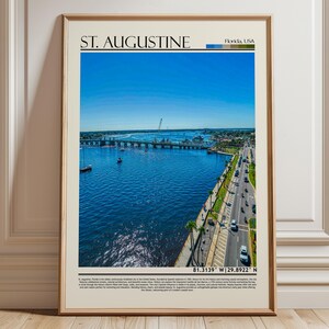 Pode incluir: Uma impressão emoldurada com uma vista aérea de St. Augustine, Flórida. A imagem mostra uma ponte sobre água azul, barcos e uma estrada com carros. O texto 'St. Augustine' está no topo, com um céu azul claro.
