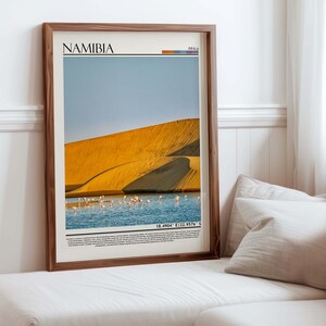 May include: Framed print featuring a scenic view of Namibia. The image showcases a golden sand dune, a body of water with pink flamingos, and a clear blue sky. The word "NAMIBIA" is printed at the top, with coordinates at the bottom.