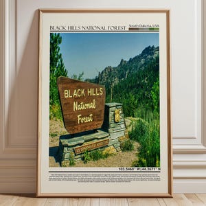 May include: A framed print featuring a photograph of the Black Hills National Forest in South Dakota, USA. The image showcases a wooden sign with the text "Black Hills National Forest" set against a backdrop of lush green trees and a mountain range.