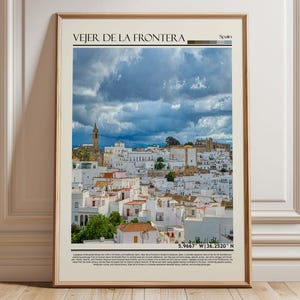 May include: Framed print of Vejer de la Frontera, Spain, featuring a cityscape of white buildings with red-tiled roofs under a dramatic cloudy sky. The print includes the town's name and geographical coordinates.