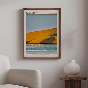 May include: A framed print featuring a Namibian landscape with a golden sand dune, blue water, and a flock of birds. The print has the word "NAMIBIA" at the top and coordinates at the bottom. The frame is made of wood.