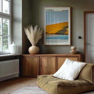 May include: A room interior featuring a framed poster of Namibia, a wooden cabinet, and a low-profile olive-green seat with a white pillow. A large vase with dried pampas grass sits atop the cabinet. A white vase is near a window.