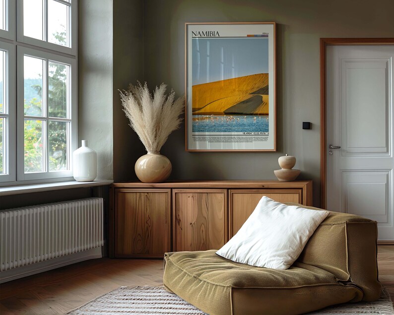 May include: A room interior featuring a framed poster of Namibia, a wooden cabinet, and a low-profile olive-green seat with a white pillow. A large vase with dried pampas grass sits atop the cabinet. A white vase is near a window.