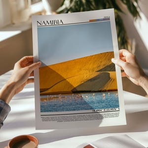 May include: A print featuring a landscape of Namibia, with a large sand dune under a blue sky. The print includes the word "NAMIBIA" at the top and coordinates at the bottom. A body of water with pink flamingos is visible.