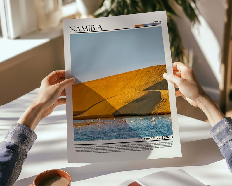 May include: A print featuring a landscape of Namibia, with a large sand dune under a blue sky. The print includes the word "NAMIBIA" at the top and coordinates at the bottom. A body of water with pink flamingos is visible.