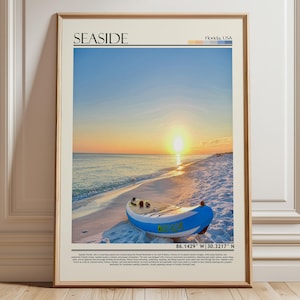 May include: Framed print of a beach at sunset, with a blue and white boat on the sand. The word "SEASIDE" is at the top. The sky changes from blue to orange as the sun sets over the sea. The print is in a wooden frame.