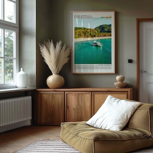 May include: A framed poster of the Philippines hangs above a wooden cabinet. A large vase with dried pampas grass sits on the cabinet. A white pillow rests on a low, olive-green cushioned seat. A white vase sits near a window.
