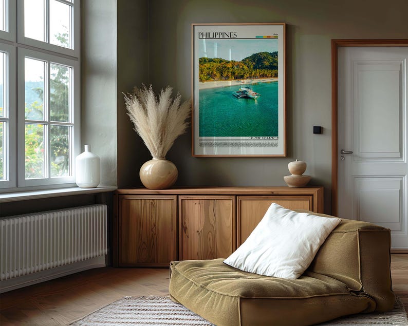 May include: A framed poster of the Philippines hangs above a wooden cabinet. A large vase with dried pampas grass sits on the cabinet. A white pillow rests on a low, olive-green cushioned seat. A white vase sits near a window.