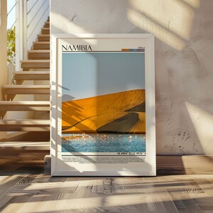 May include: A framed print featuring a scenic view of Namibia. The artwork showcases a golden sand dune, a body of water, and a blue sky. The word "NAMIBIA" is printed at the top. The print is displayed on a wooden floor, leaning against a textured wall.