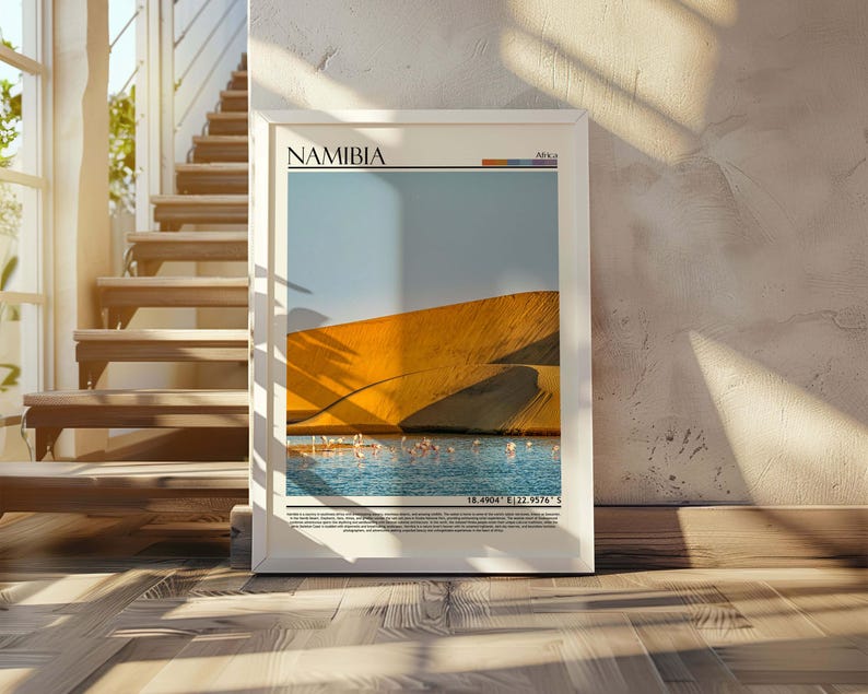 May include: A framed print featuring a scenic view of Namibia. The artwork showcases a golden sand dune, a body of water, and a blue sky. The word "NAMIBIA" is printed at the top. The print is displayed on a wooden floor, leaning against a textured wall.