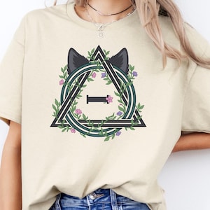 May include: A sand-colored short-sleeve t-shirt featuring a graphic print of a black triangle framing a circular design with green vines, small purple flowers, and dark animal ears. The center of the design contains the Greek letter Theta.