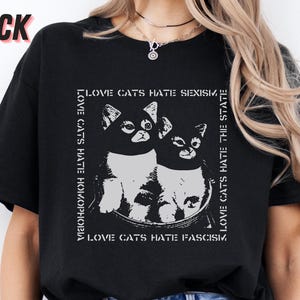 Op de afbeelding: Zwart T-shirt met een afbeelding van twee katten in een kom en de tekst "LOVE CATS HATE SEXISM, HOMOPHOBIA, FASCISM, THE STATE." Het woord "BLACK" staat linksboven. Het shirt is gemaakt van zacht materiaal.