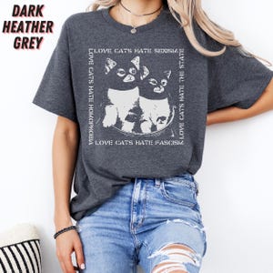 Op de afbeelding: Donker heather grijs t-shirt met een witte afbeelding van twee katten en de tekst "LOVE CATS HATE HOMOPHOBIA, SEXISM, FASCISM, THE STATE". Het shirt heeft een ronde hals en korte mouwen.