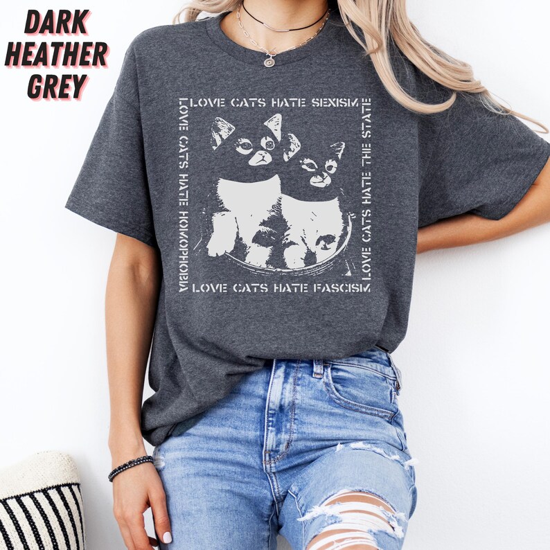 Op de afbeelding: Donker heather grijs t-shirt met een witte afbeelding van twee katten en de tekst "LOVE CATS HATE HOMOPHOBIA, SEXISM, FASCISM, THE STATE". Het shirt heeft een ronde hals en korte mouwen.