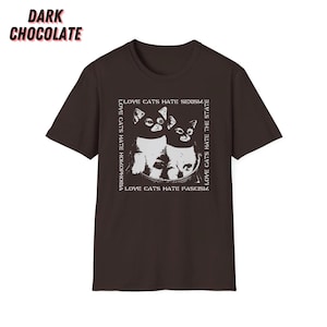 Op de afbeelding: Donkerchocoladekleurig T-shirt met een witte afbeelding van twee katten en de tekst: "LOVE CATS HATE HOMOPHOBIA, SEXISM, FASCISM, THE STATE". De tekst en de kattenafbeelding zijn wit.