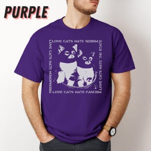 Op de afbeelding: Paars T-shirt met een witte afbeelding van twee katten en de tekst "LOVE CATS HATE SEXISM, HOMOPHOBIA, THE STATE, FASCISM". Het woord "PURPLE" staat in grote, vette letters linksboven.