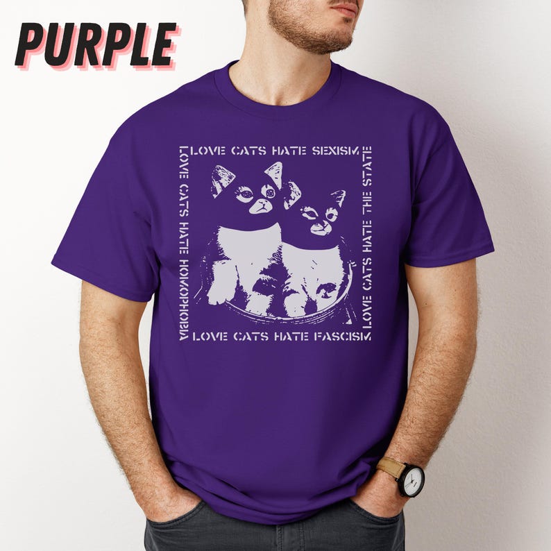 Op de afbeelding: Paars T-shirt met een witte afbeelding van twee katten en de tekst "LOVE CATS HATE SEXISM, HOMOPHOBIA, THE STATE, FASCISM". Het woord "PURPLE" staat in grote, vette letters linksboven.