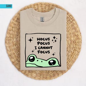 Può includere: Una felpa girocollo color sabbia con una grafica quadrata dal bordo nero. All'interno del quadrato, il testo recita HOCUS POCUS I CANNOT FOCUS, accompagnato da piccole icone di stelle nere e un'illustrazione di rana verde chiaro.