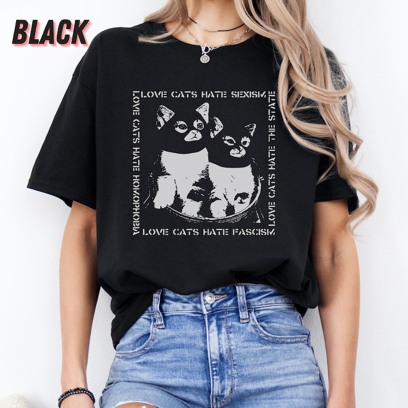 Op de afbeelding: Zwart T-shirt met een afbeelding van twee katten in een kom en de tekst "LOVE CATS HATE SEXISM, HOMOPHOBIA, FASCISM, THE STATE." Het woord "BLACK" staat linksboven. Het shirt is gemaakt van zacht materiaal.