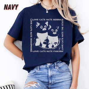 Op de afbeelding: Marineblauw T-shirt met een witte afbeelding van twee katten in een vierkant kader. De tekst rond de katten luidt "LOVE CATS HATE SEXISM", "LOVE CATS HATE HOMOPHOBIA", "LOVE CATS HATE THE STATE" en "LOVE CATS HATE FASCISM".