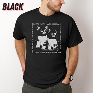 Op de afbeelding: Zwart t-shirt met een witte afbeelding van twee katten en de tekst "LOVE CATS HATE HOMOPHOBIA, SEXISM, THE STATE, FASCISM." Het woord "BLACK" staat linksboven gedrukt. Het ontwerp is wit.