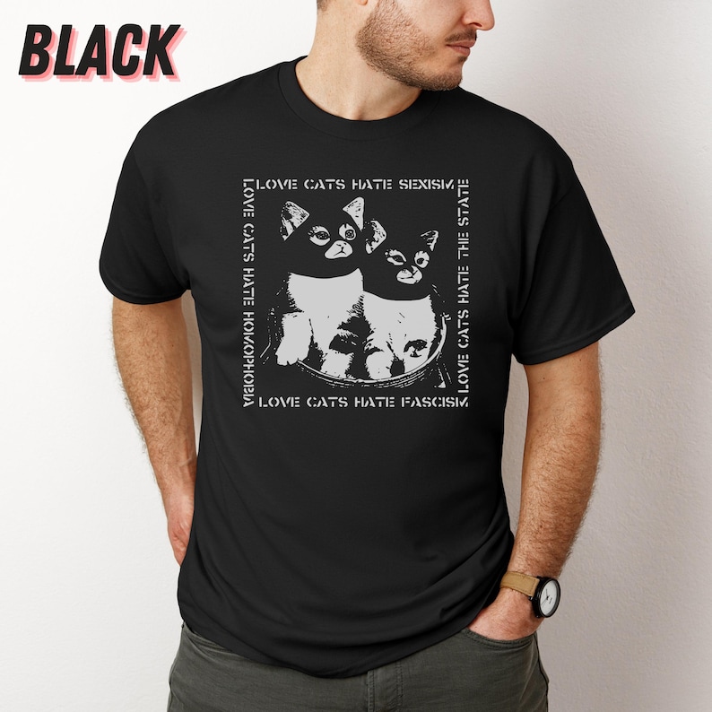 Op de afbeelding: Zwart t-shirt met een witte afbeelding van twee katten en de tekst "LOVE CATS HATE HOMOPHOBIA, SEXISM, THE STATE, FASCISM." Het woord "BLACK" staat linksboven gedrukt. Het ontwerp is wit.