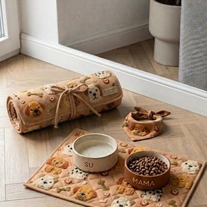 Peut inclure: Un tapis de compagnie matelassé beige avec des visages de chiens et des os brodés, un tapis roulé avec des attaches, et un petit bandana. Deux bols en céramique sont posés sur le tapis, l'un marqué SU et l'autre MAMA, contenant de l'eau et des croquettes.