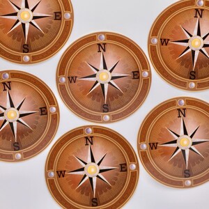 Puede incluir: Seis posavasos marrones en forma de brújula con un borde dorado y un diseño de rosa de los vientos blanco. Cada posavasos tiene las letras N, S, E y W alrededor de la rosa de los vientos.