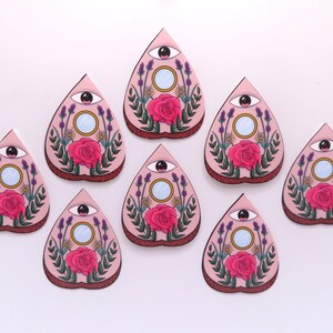 May include: A collection of eight pink, teardrop-shaped decorative items. Each features a central rose, an eye, a mirror, and floral accents. The items are arranged on a white surface, creating a visually appealing pattern.