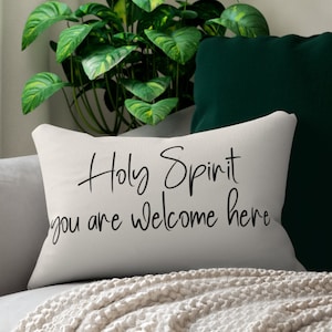 May include: White throw pillow with black text that reads "Holy Spirit you are welcome here". The pillow is on a couch with a green pillow and a white blanket.