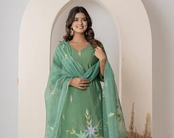Puede incluir: Un conjunto salwar kameez verde bordado. El atuendo incluye una túnica, pantalones y una bufanda transparente. La túnica presenta bordados florales en tonos verdes y blancos. El conjunto está diseñado con un escote en V y está hecho de una tela suave y fluida.