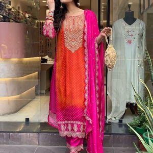 May include: A vibrant orange and hot pink salwar kameez featuring a dotted pattern, intricate gold embroidery on the chest and hem, and a matching hot pink dupatta with a scalloped border. A small, beaded, gold-toned drawstring pouch is held.