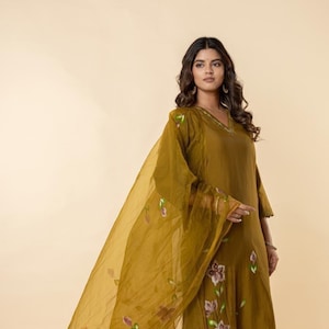 May include: Olive green tunic and matching sheer scarf with floral accents. The tunic has a V-neck and three-quarter sleeves. The scarf drapes over the shoulder and features painted floral designs. The outfit is a flowing, elegant style.